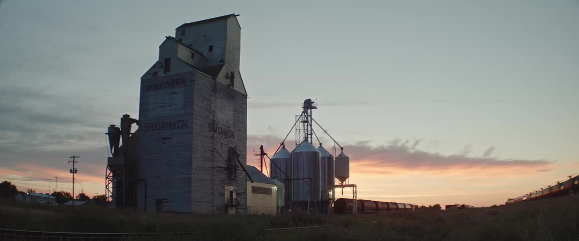 A grain elevator with silos sits near railroad cars at sunrise