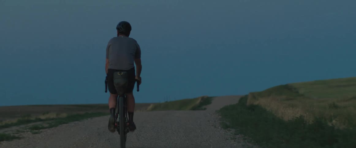 Man wearing a helmet rides a bicycle on a rural dirt road at dusk
