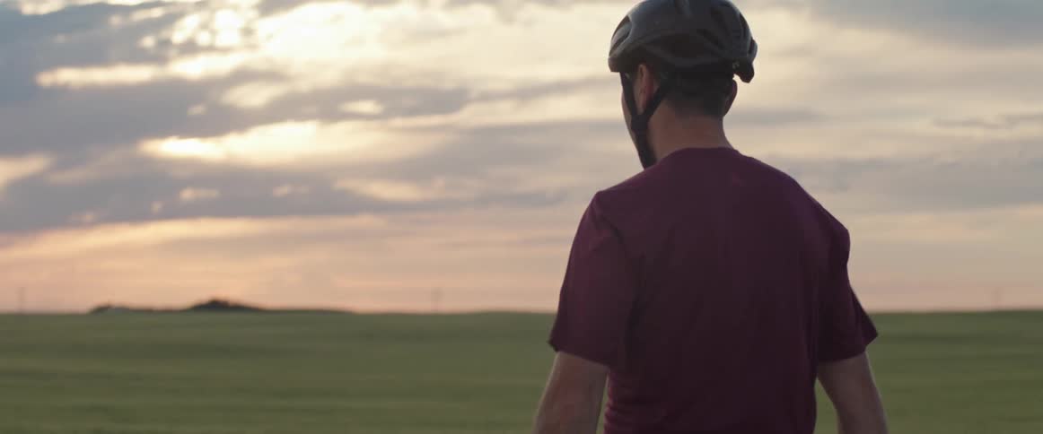 Man wearing a bike helmet riding his bike down a dirt road next to a grass field during sunset