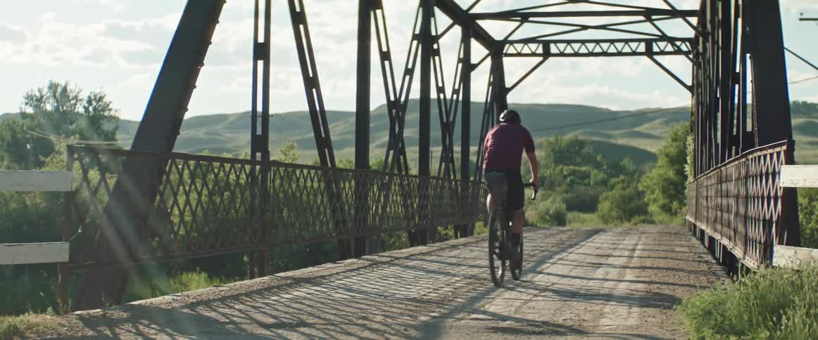 A man is riding a bicycle across a bridge and there are mountains in the distance under a sunny sky