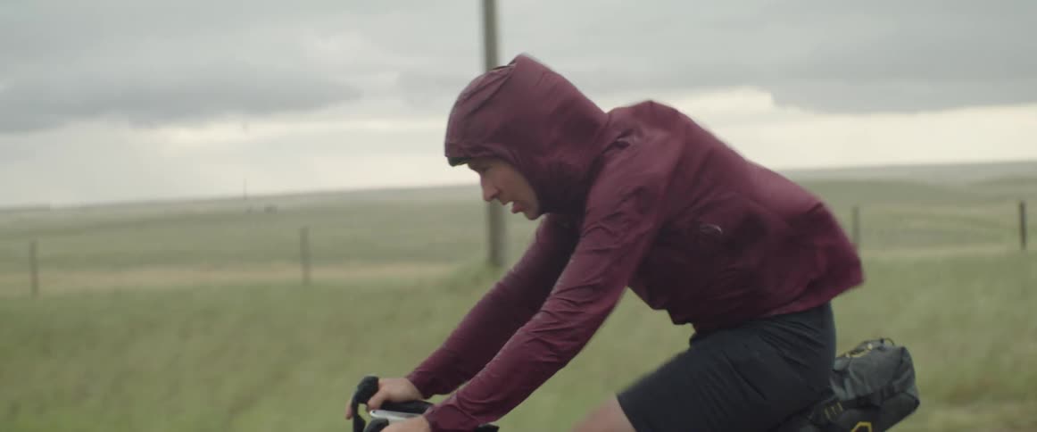 Man wearing a hooded jacket riding a bicycle during a windy day on a dirt road