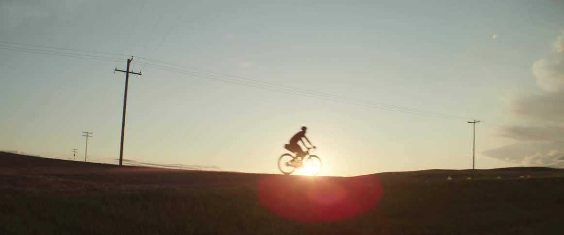 Silhouette of a man riding a bicycle on a road with utility poles and the sun shining brightly on the horizon