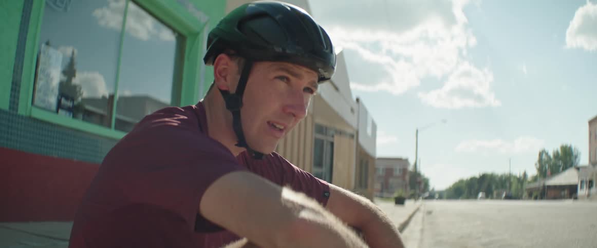 Man with black helmet and red t shirt sits on side walk sweating in the town square
