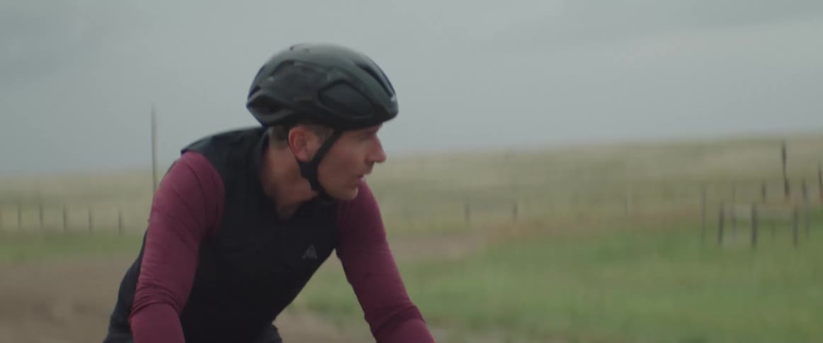 A man wearing a helmet is riding a bicycle on a rural road under a cloudy sky