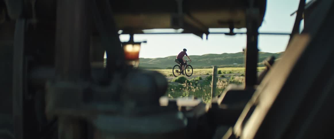 A man rides his bike on a country road past farm equipment and green hills