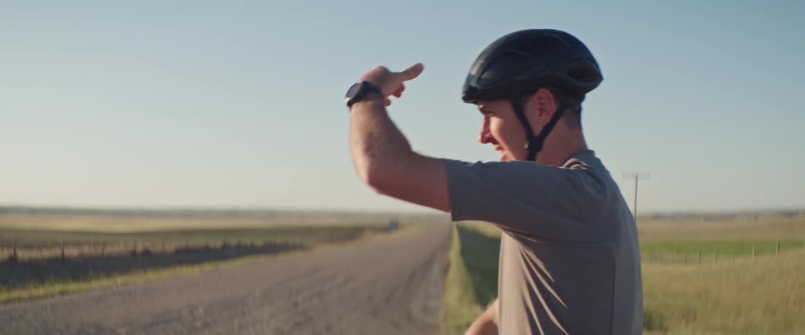 Man wearing a helmet and riding a bike stopping near a dirt road and looking around while covering his face against the sunlight with his hands