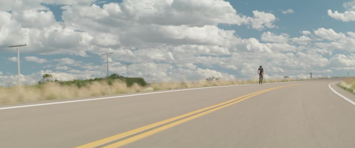 A man is riding a bicycle on a rural road under a big white clouds