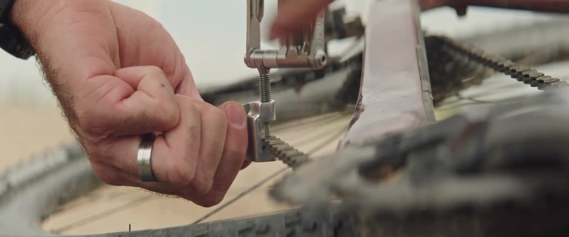Man works on bicycle chain with hand tool while the bicycle sits on its side on the ground