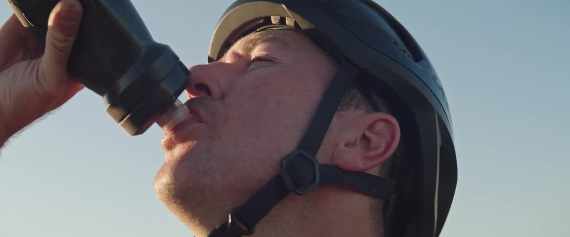 Man wearing bicycle helmet rests on road in countryside and drinks water from a water bottle