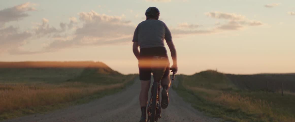 A man is riding a bicycle and pedalling hard on a wide open rural dirt road during sunset