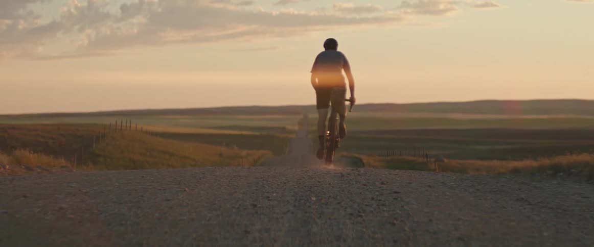 Male cyclist rides bicycle over crest of country road at sunset