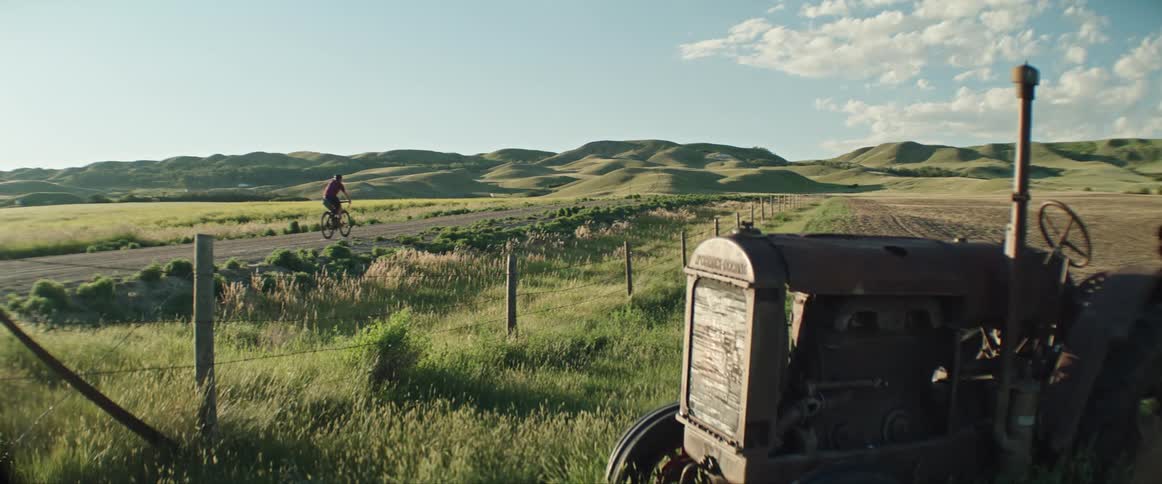 Cyclists pedals along road past crop field with old parked tractor