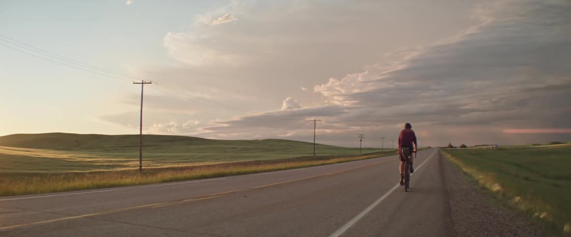 Man riding a bicycle on an empty road towards storm clouds in the setting sun