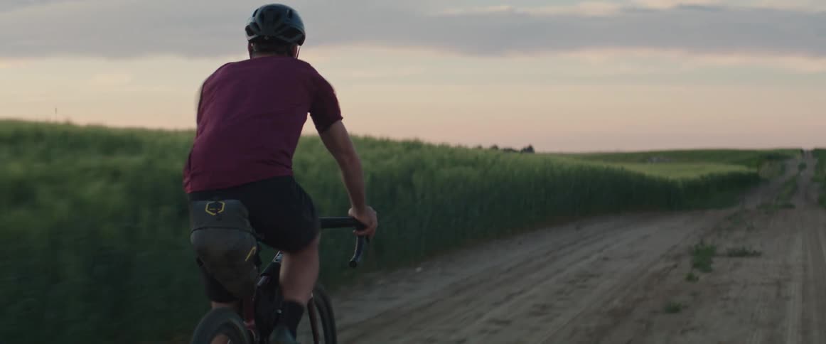 Male cyclist with helmet red shirt and black shorts rides bicycle on country road next to field during sunset