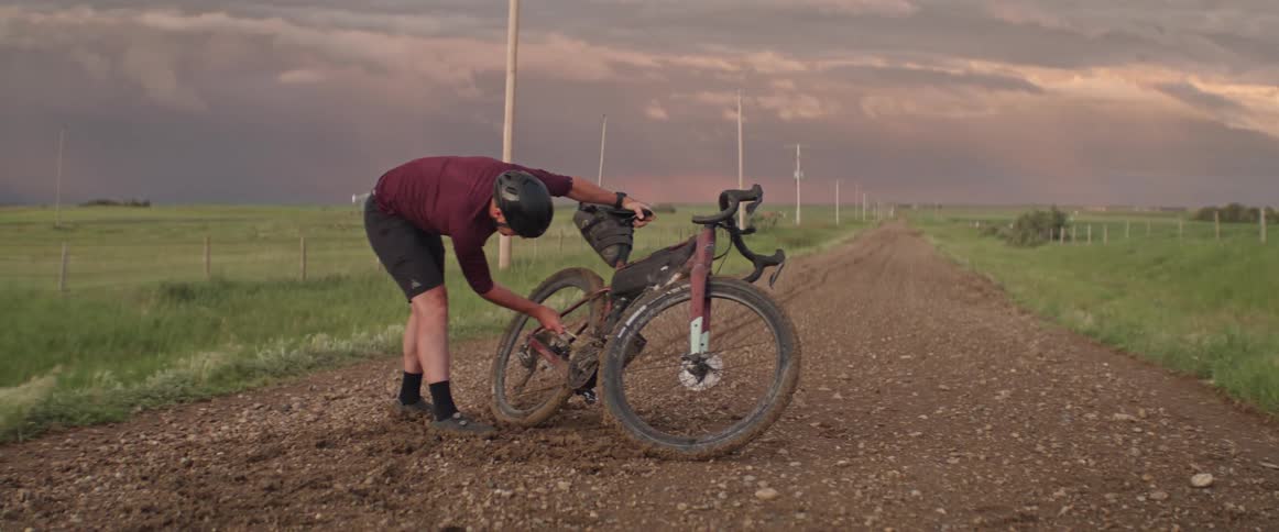 Man wearing a bike helmet trying to fix his bicycle in the middle of a dirt road