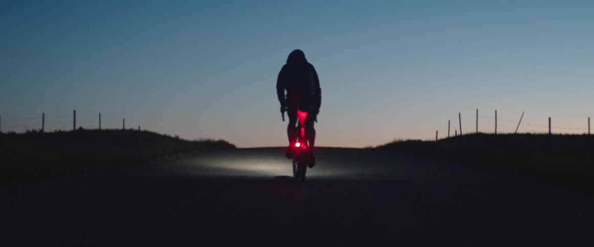 Cyclist approaches the top of a rural road hill while silhouetted against the dusk sky