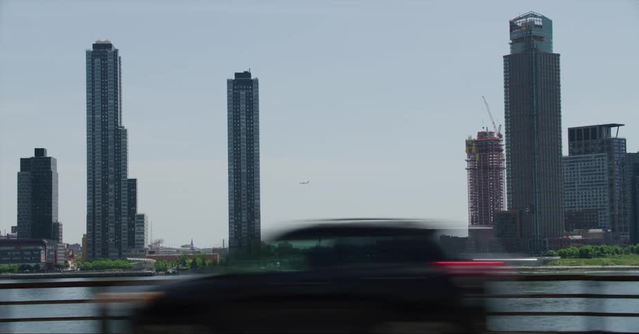 Vehicles drive along a road that is across a river from several tall skyscrapers