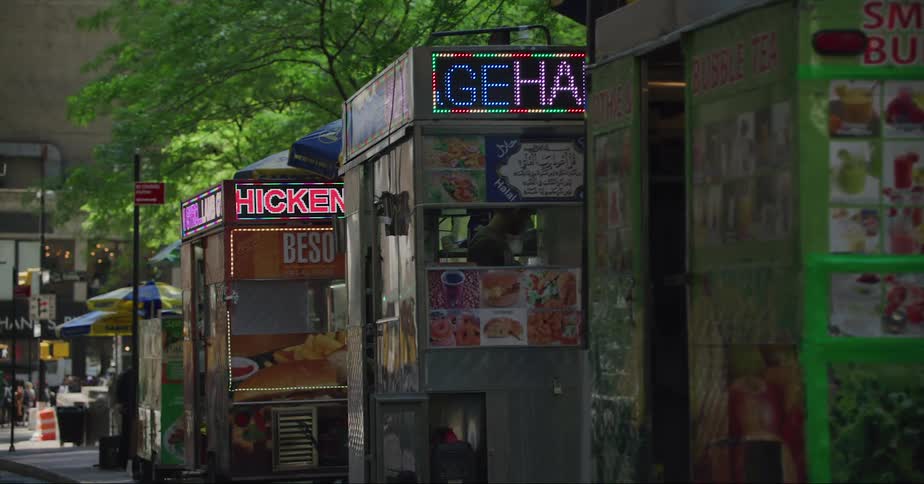 Stands and booths are set up on the street with lights and signs with trees and buildings in the distance