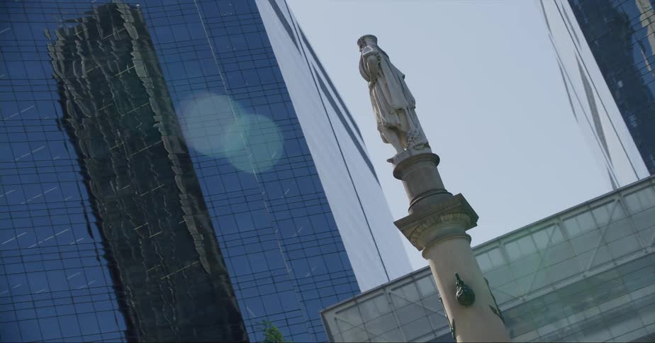 A statue stands atop a tall pedestal amidst several tall buildings