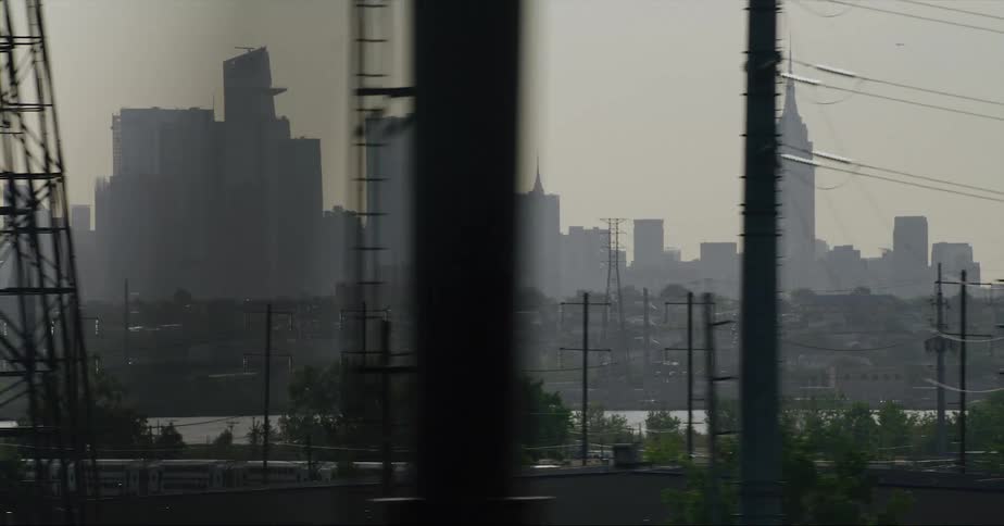Tall city buildings in a hazy sky are seen from a distance as a train moves along the harbor