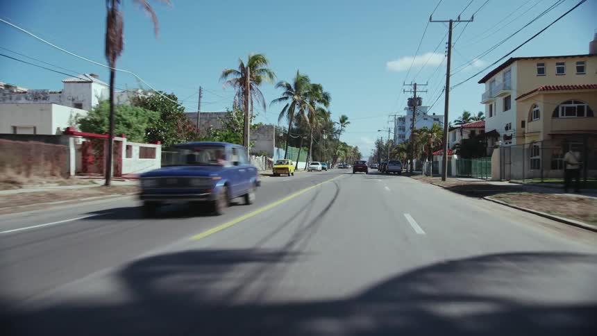 Traffic moves on a street with automobiles parked on the roadside lined with palm trees and buildings