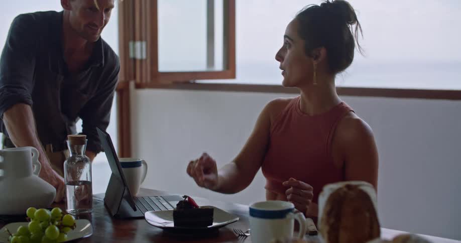 A woman sitting at a table with her laptop discusses and points at a piece of cake