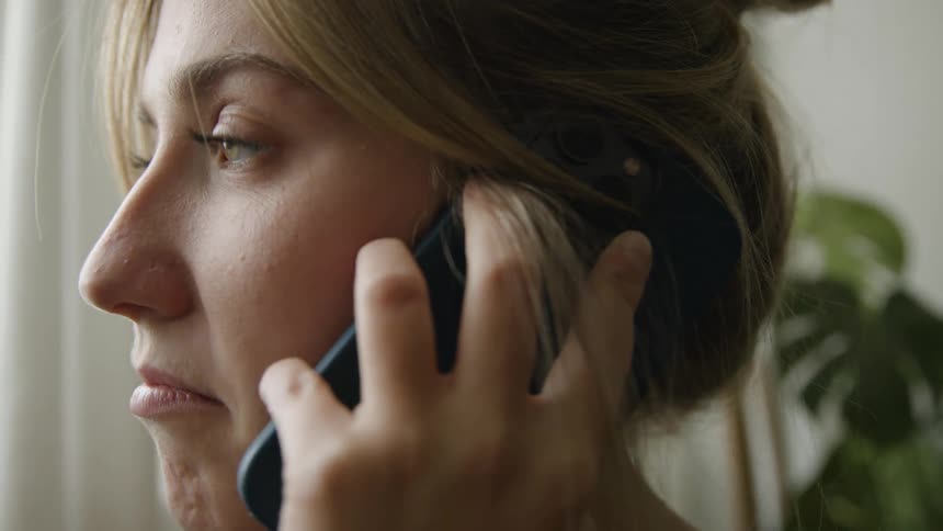 A woman is inside her home holding her phone to her face as she talks