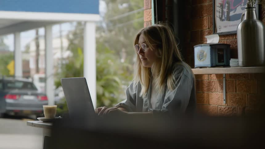 A woman puts on her eye glasses and smiles as she types while sitting in a coffee shop