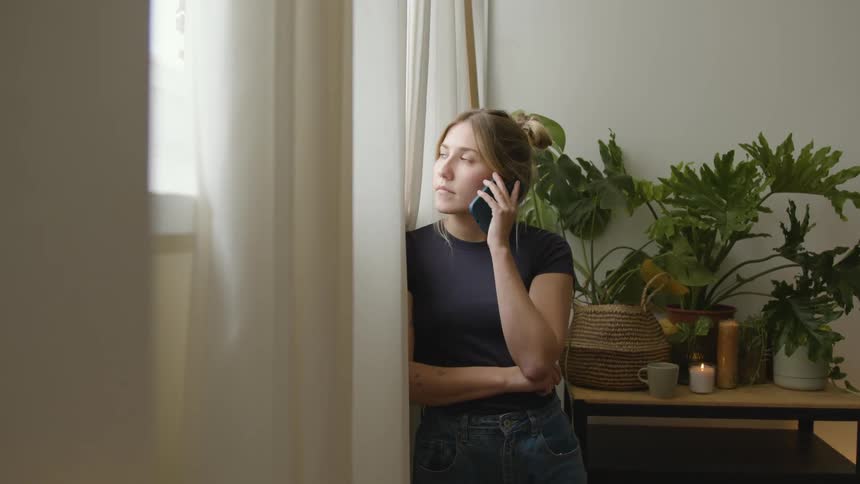 A woman stands next to the window while making a phone call