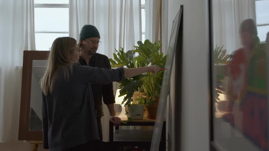 Man with gray beanie glasses and dark shirt stands next to blonde woman discussing in front of whiteboard in room