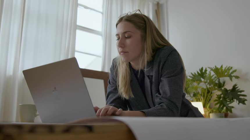 A woman leans against her desk while typing on her laptop