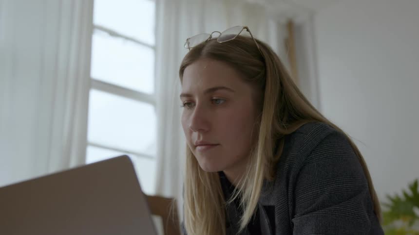 A woman is sitting in front a laptop computer and touches her pen to her mouth as she smiles and looks at the screen
