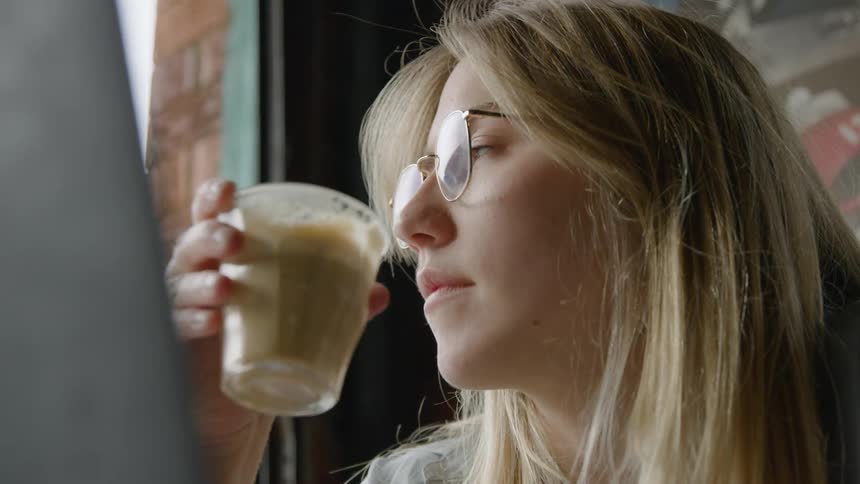 Blonde woman with glasses drinks coffee from glass sitting in front of laptop in cafe