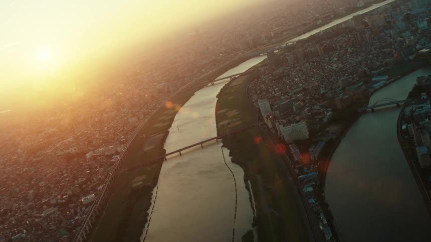 Bridges cross a river outside a big city with tall buildings as the sun shines brightly in a hazy sky