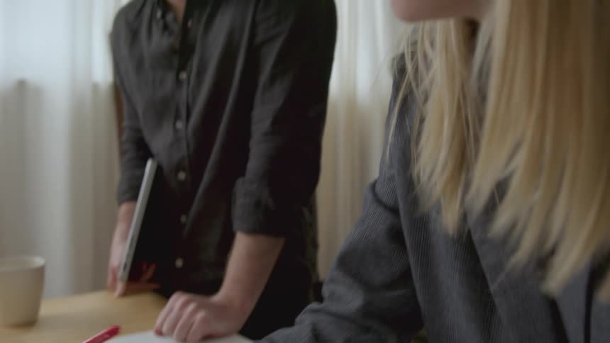 A woman is sitting at a table drawing plans with a ruler as she talks to a man standing next to the table
