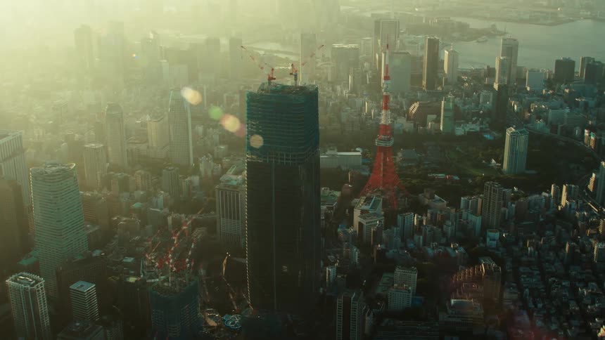An aerial view of construction cranes on a high rise building in a city filled with skyscrapers and a river in the distance