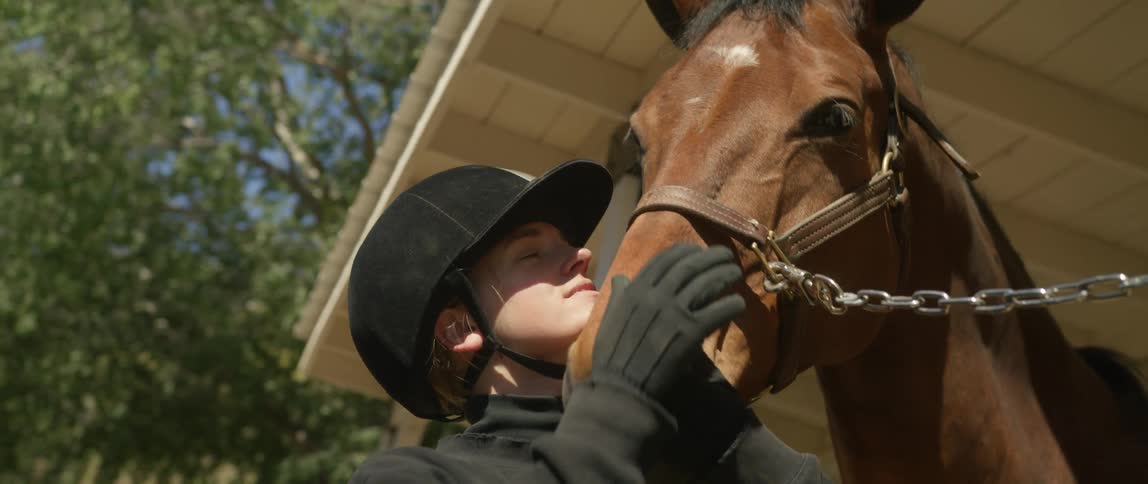 Young female jockey caresses and kisses the snout of a horse tied outside a stable