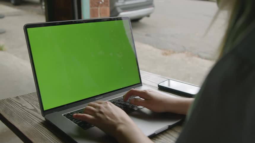 A woman is outside sitting at a table typing on a laptop computer with a green screen