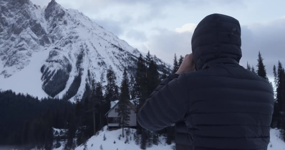 Person in winter coat stands on a hill looking at snow covered house and mountain