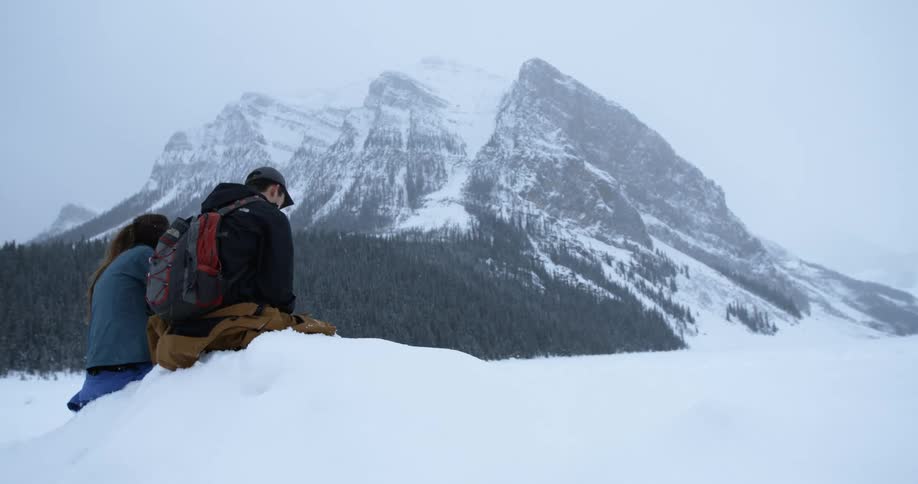 A man and women sit on some snow looking over towards a mountain