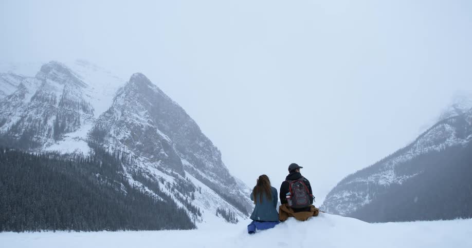 A man and woman sit on a snow pile while it snows in the mountains