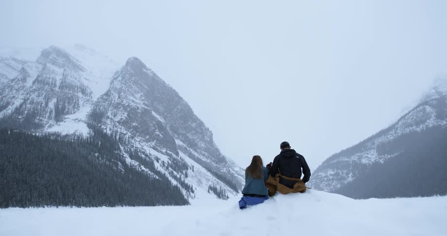 A man and woman sit on a snowbank and look at mountains