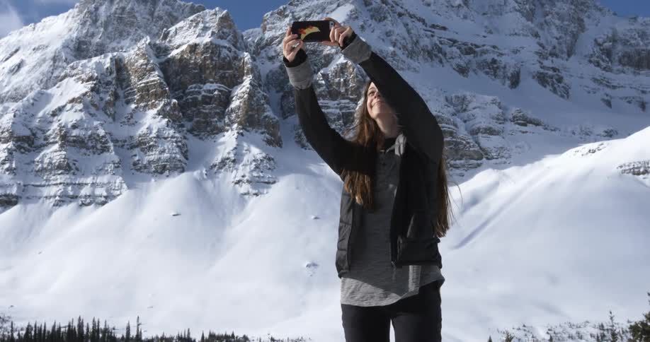 A woman is raising her phone in the air before a snow covered mountain
