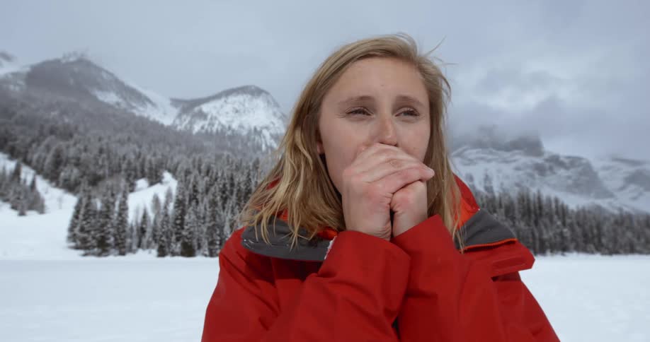 A woman warms her hands by blowing on them in a snowy opening in the mountains
