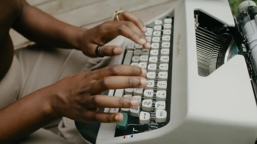 Woman wearing rings beige pants and black top types on typewriter