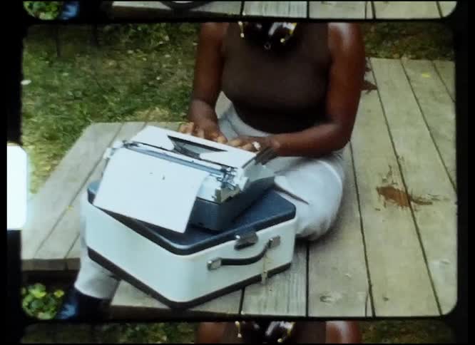 A woman smiles as she sits on a wooden deck in the backyard typing on a typewriter