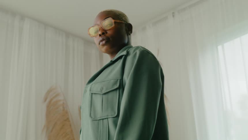 Woman with short hair sunglasses and green jacket poses for portrait in room with curtains