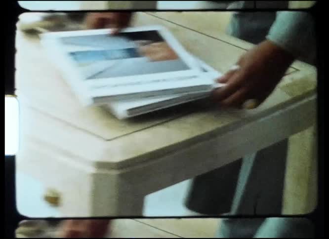Woman with short hair sunglasses and green jacket puts books on coffee table in front of yellow couch in living room