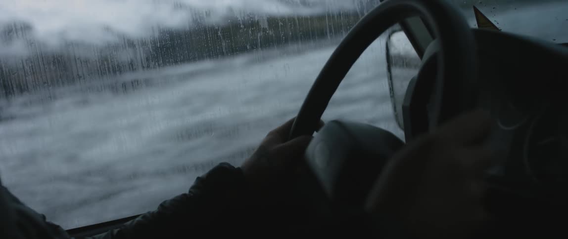 A man drives his vehicle through shallow river water past snow capped mountains