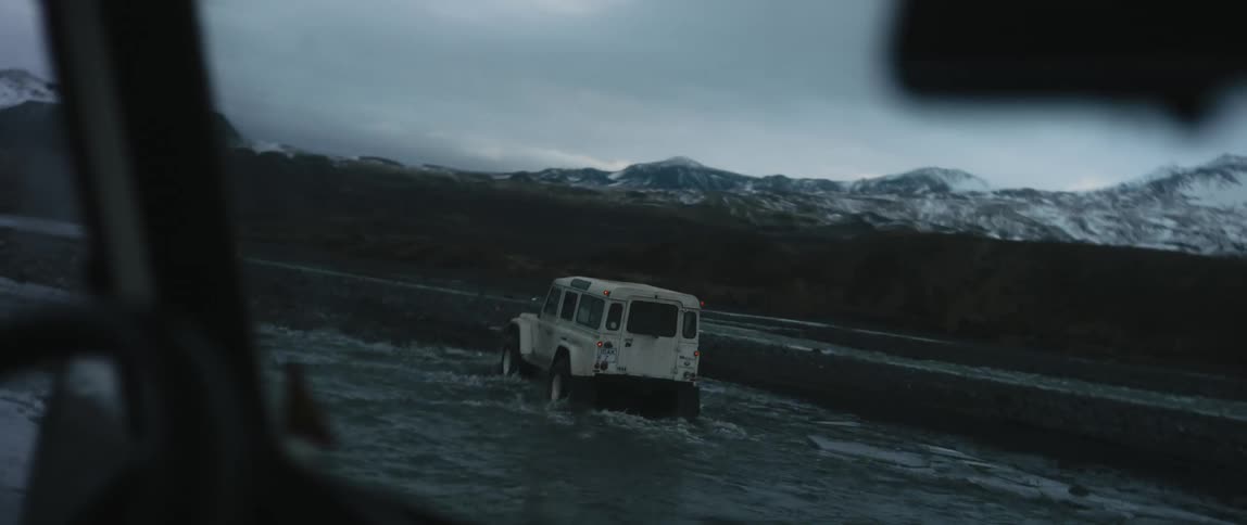 A group of offroad vehicles travel across a shallow stream in the wilderness during the day